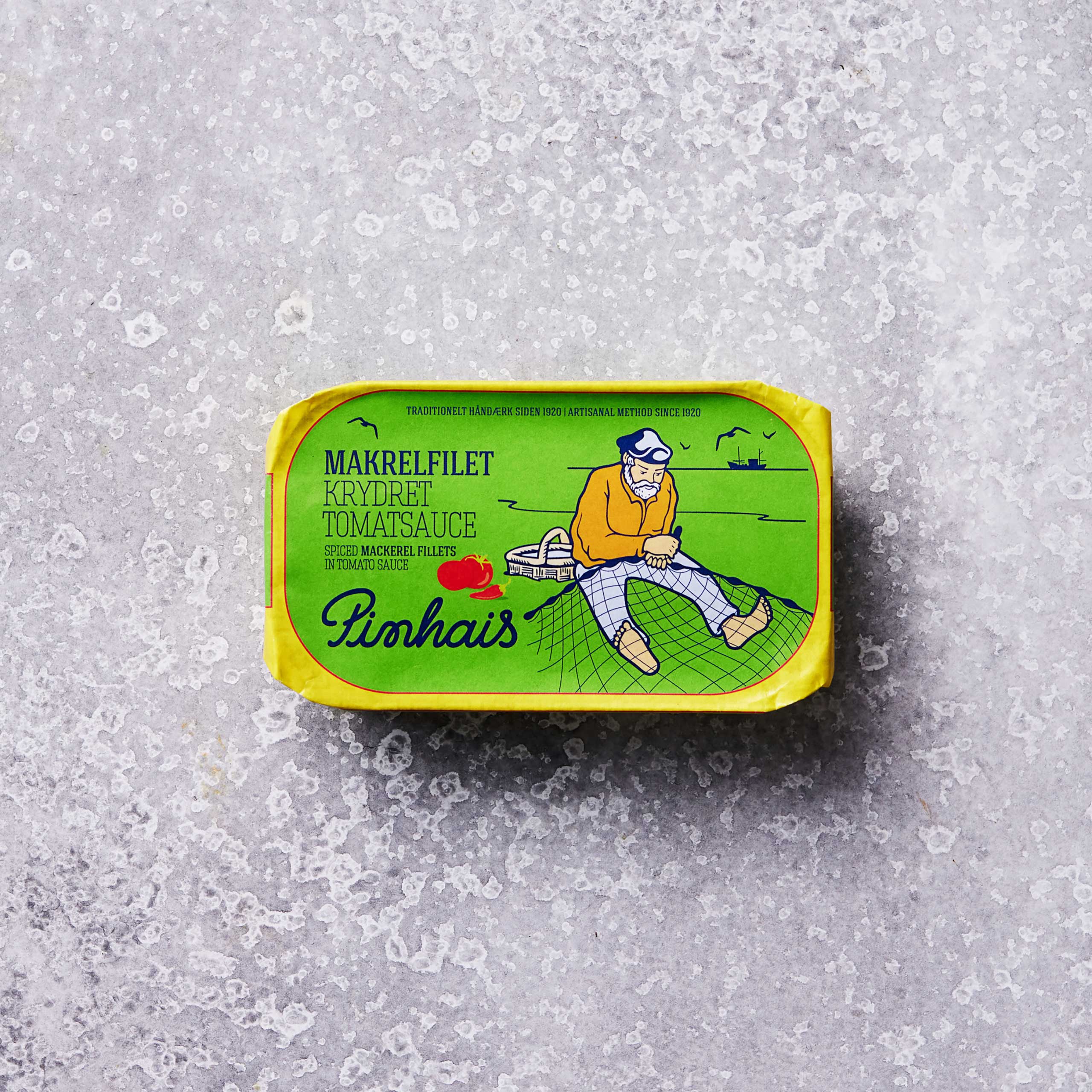 En dåse Pinhais Makrelfilet i krydret tomatsauce med en grøn etiket, der viser en skægget mand i gult på et fiskenet og en båd i baggrunden.