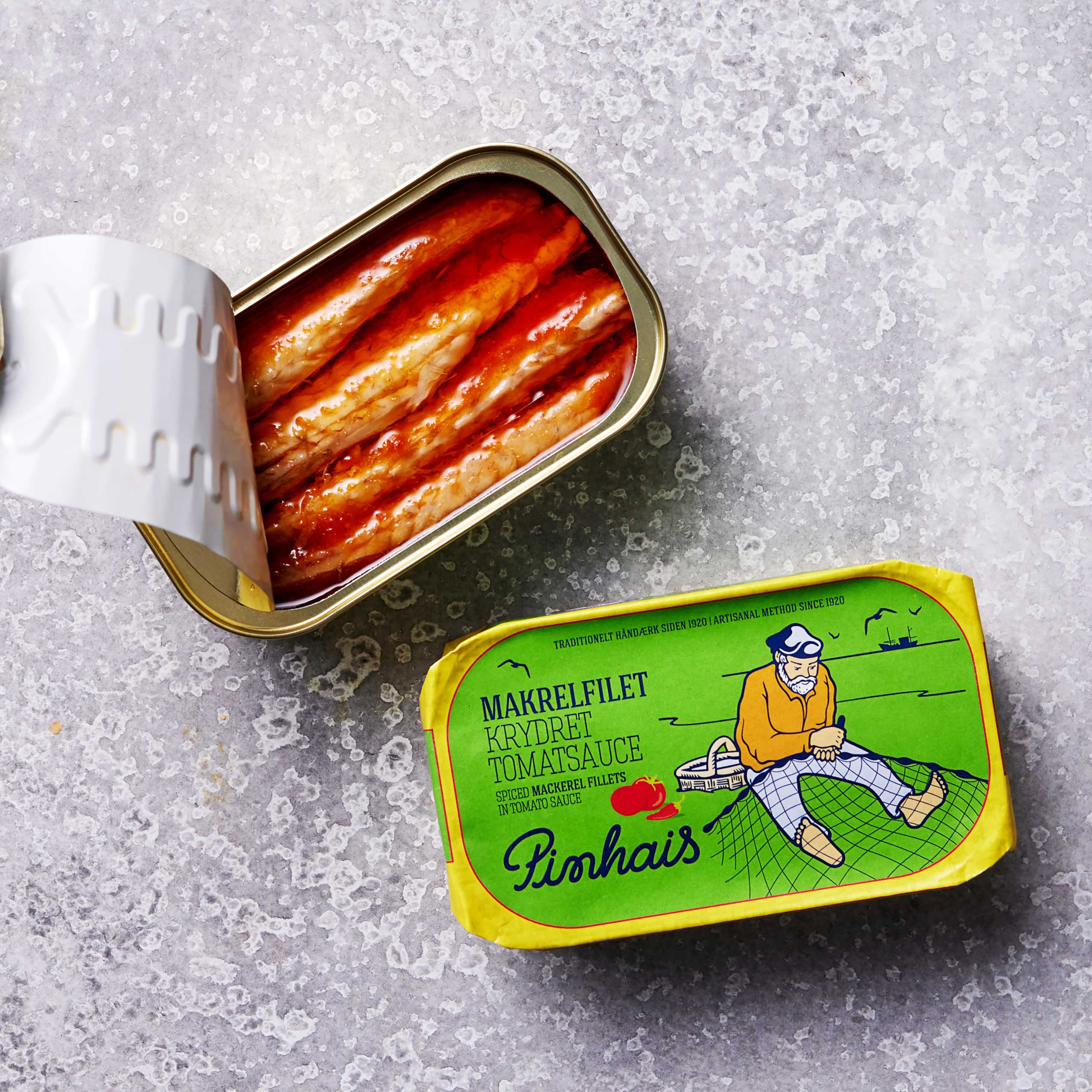 En åbnet dåse Pinhais Makrelfilet i krydret tomatsauce sidder på en grå overflade ved siden af dens gule og grønne emballage, med en mand, der fisker - perfekt til dem, der søger omega-3 fedtsyrer fra bæredygtigt fangede makreller.