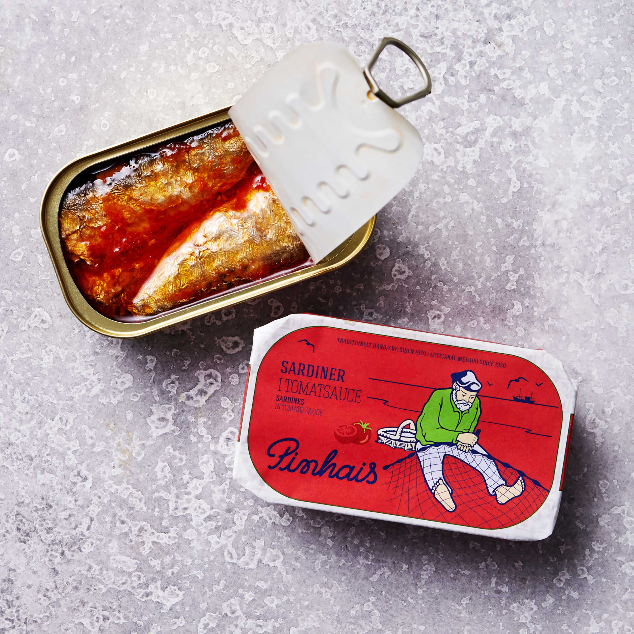 En åbnet dåse Portugisiske sardiner i tomat og olivenolie sidder ved sin emballage, som viser en fisker i en grøn sweater på et rødt net, med Pinhais-mærke i rødt og blåt. Rik på omega-3-fedtsyrer og solmodne tomater.