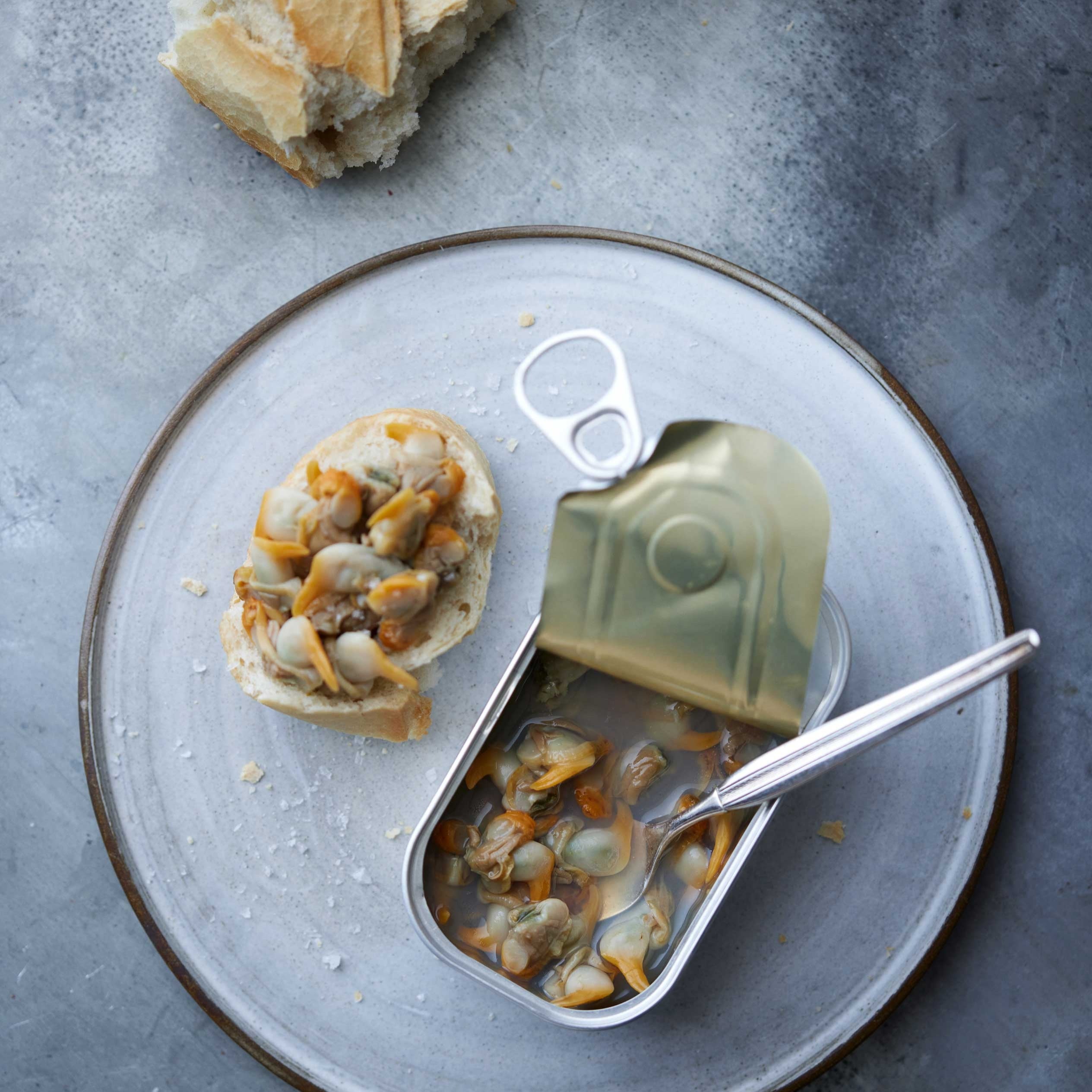 En tallerken holder en skive brød toppet med "Don Gastronom Hjertemuslinger i egen lage" ved siden af en åben dåse med muslinger i lage og en gaffel. Et stykke brød hviler på tallerkenens kant, helt nede på en grå overflade. Bæredygtig Galicien skaldyr.