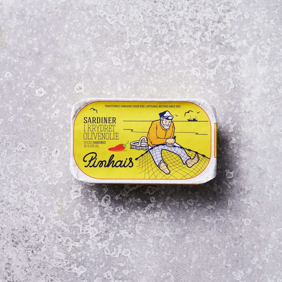 En rektangulær dåse med bæredygtige sardiner har en gul etiket med en illustration af en mand, der sidder på et fiskenet, og den norske tekst "Sardiner i krydret olivenolie" og mærkenavnet "Pinhais" på forsiden.