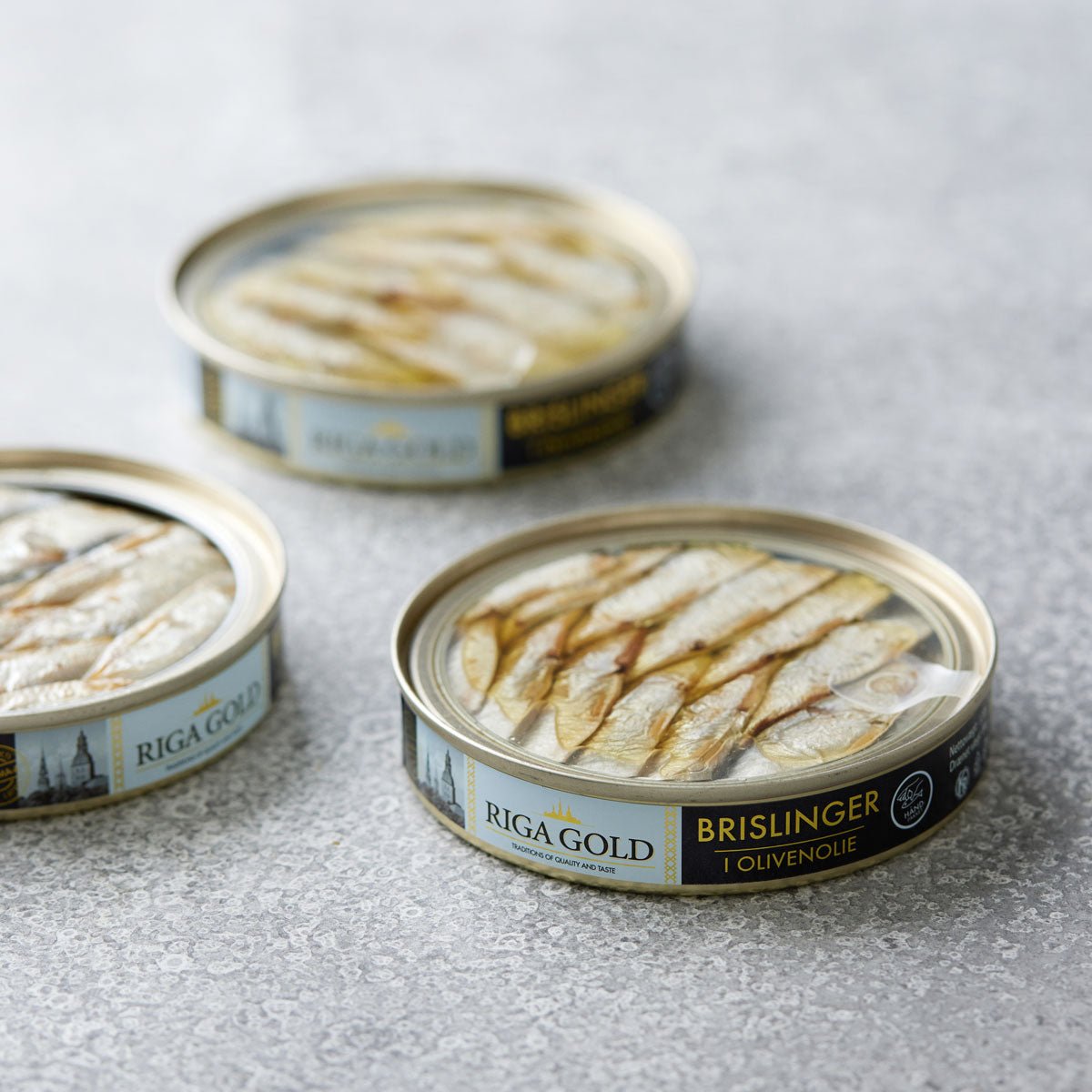 Tre åbne dåser Riga Gold Små brislinger i olivenolie er arrangeret på en tekstureret lysegrå overflade, med fisken pænt pakket og etiketterne synlige – en sand delikatesse blandt nordiske sardiner.