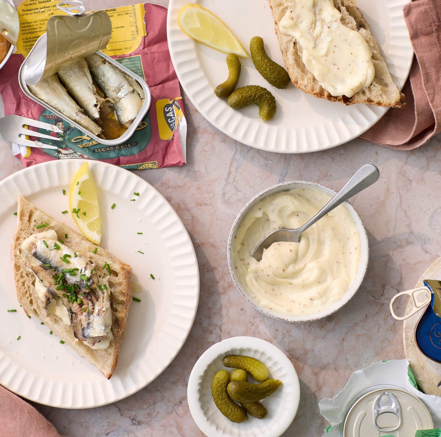 To tallerkener med sardiner på baguetteskiver, toppet med Grøndals Mayonnaise – perfekt til fisk og purløg, står ved siden af en skål med sauce, citronbåde, syltede agurker og en åben dåse Grøndals fiskekonserves på et lyst marmorbord.
