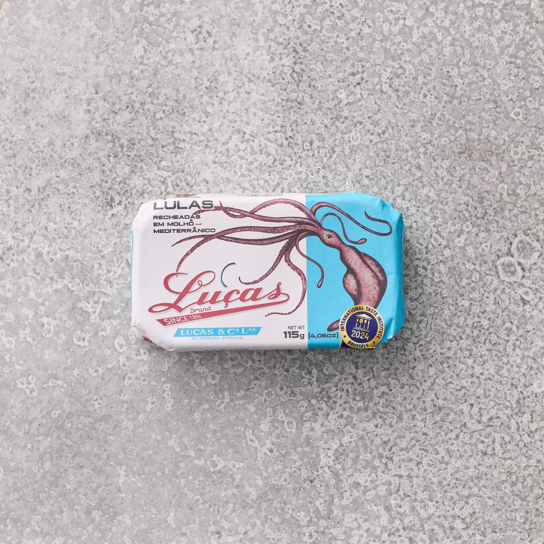 En rektangulær dåse Lucas Fyldte blæksprutte i middelhavssauce med en blæksprutteillustration på den blå og hvide etiket ligger på en tekstureret grå overflade og fremkalder klassiske middelhavsblækspruttesmag.