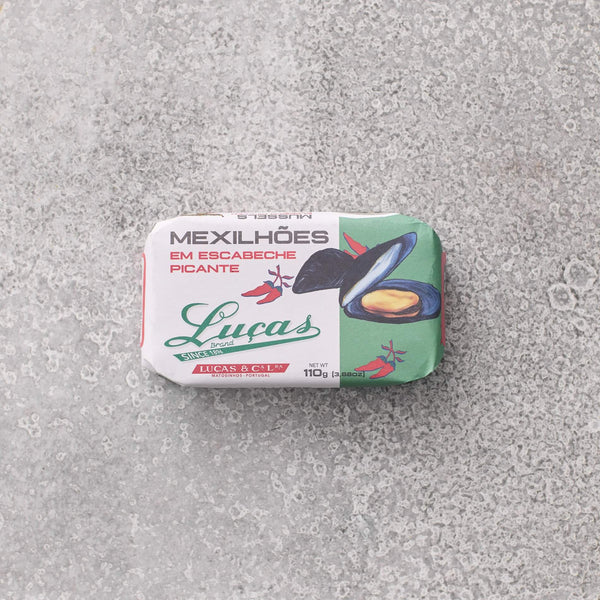 En dåse Lucas Muslinger i krydret escabeche-sauce med en illustration af en åben muslingerskal og portugisisk tekst ligger på en grå tekstureret overflade.