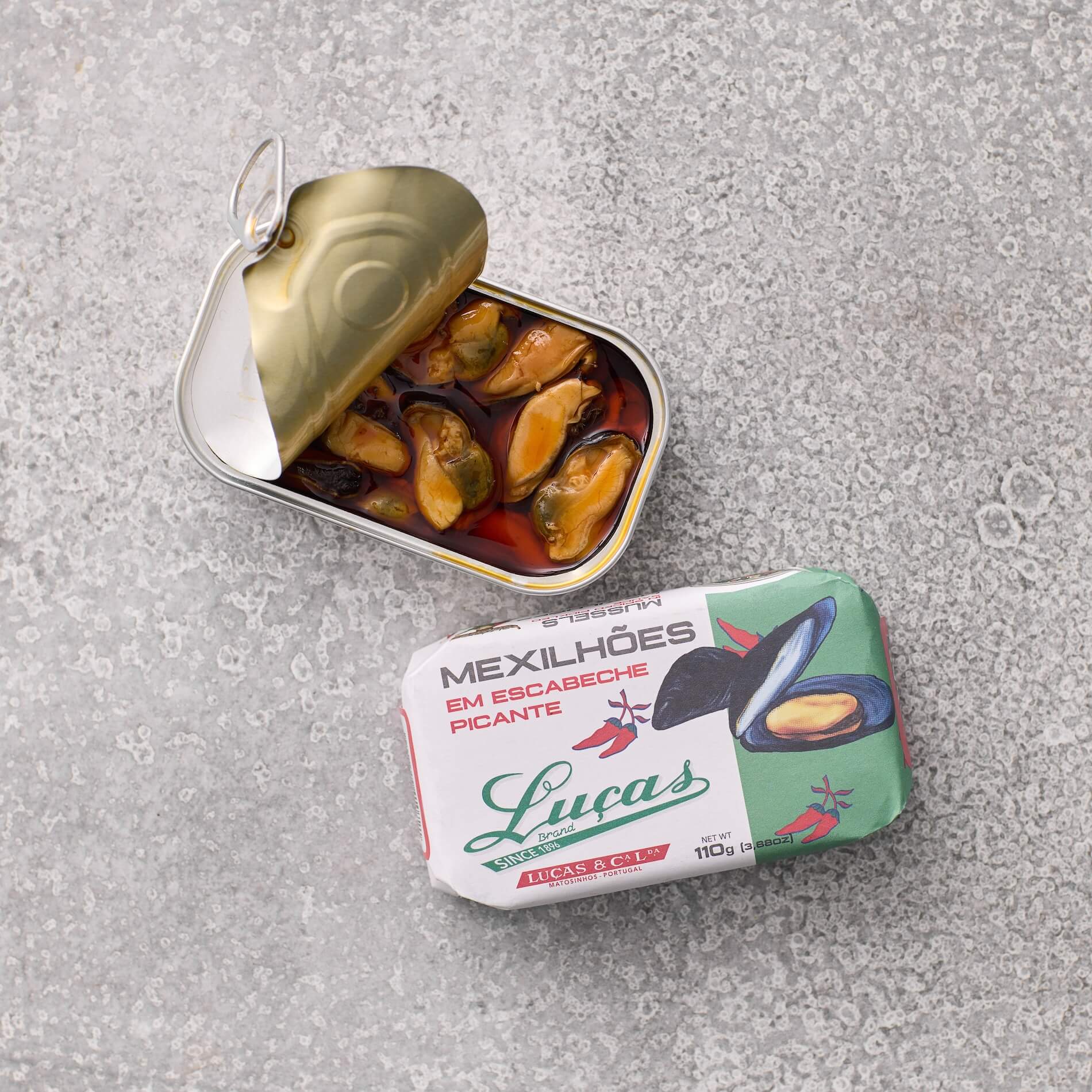 En åbnet dåse Muslinger i krydret escabeche-sauce fra Lucas ligger ved siden af emballagen, som viser en illustration af muslinger og Lucas-mærket på en grå tekstureret overflade.