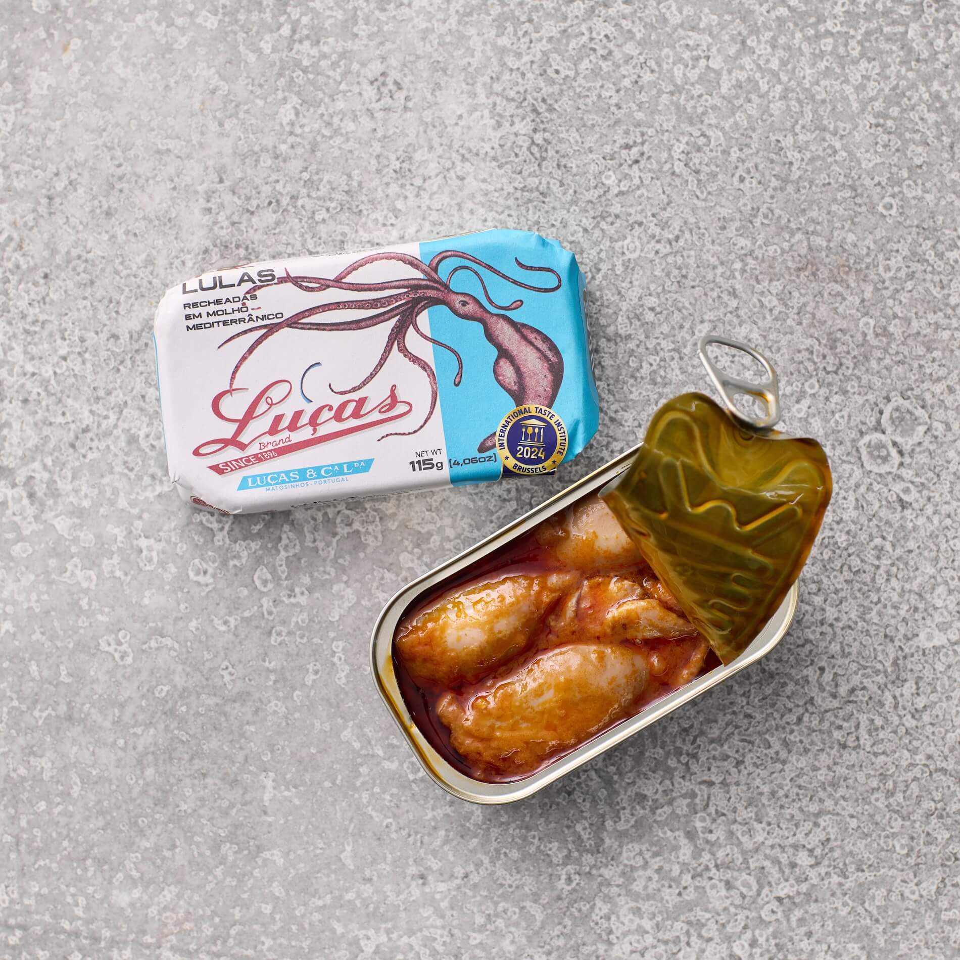 En åben dåse Lucas Fyldte blæksprutter i middelhavssauce ligger på en grå tekstureret overflade ved siden af den lukkede emballage, som viser en illustration af blæksprutte og Lucas-mærket.