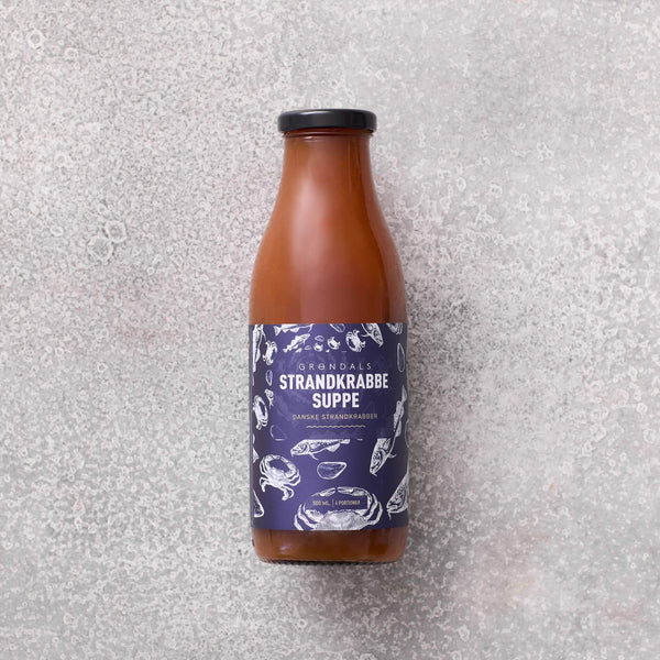 En glasflaske fyldt med brun suppe med en mørkeblå etiket med teksten Grøndals Strandkrabbe Suppe og hvide illustrationer af krabber, skaller og skaldyr, placeret på en lysegrå struktureret overflade.