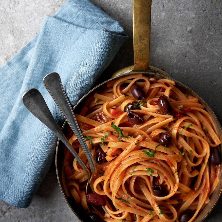 En gryde med linguine-pasta i tomatsauce med sorte oliven, pyntet med hakket persille. To metalgafler hviler i gryden, som står ved siden af en foldet lyseblå stofserviet på en stenoverflade.