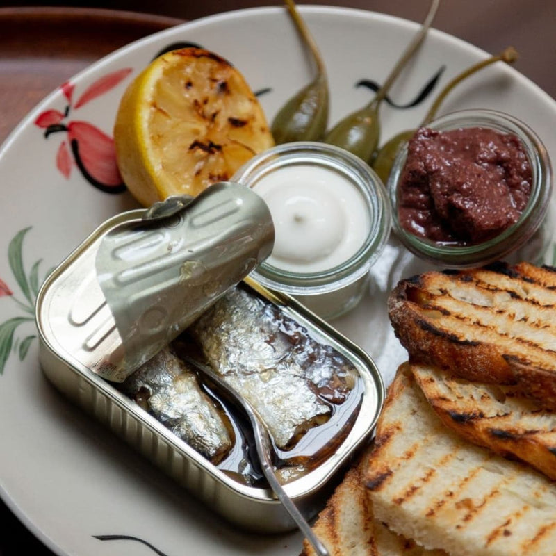 En tallerken med en åben dåse sardiner, grillet citron, kapersbær, to små glas med saucer eller smørepålæg og skiver af ristet brød. Maden er arrangeret pænt på en dekorativ tallerken.