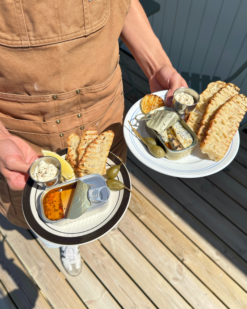 En person i brunt forklæde står på et solbeskinnet trædæk med to tallerkener med fiskekonserves, fladbrød, kapersbær, en citronbåd, pålæg og kiks.