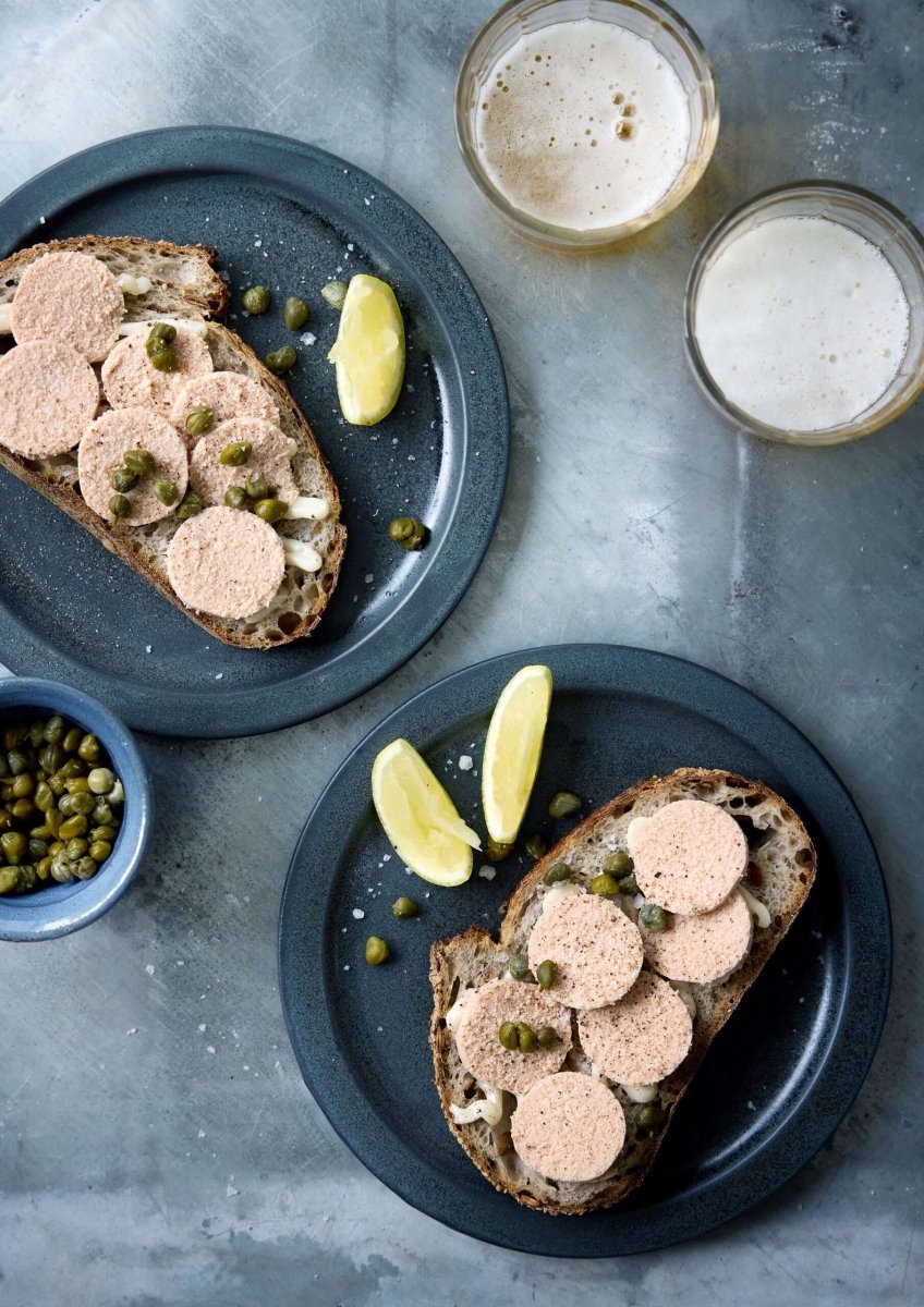 To tallerkener med hver en skive brød toppet med runde skiver af Grøndals Islandsk torskerogn fra vildtfanget torsk og kapers, serveret med citronbåde. To glas letøl og en skål med kapers fuldender bordet.