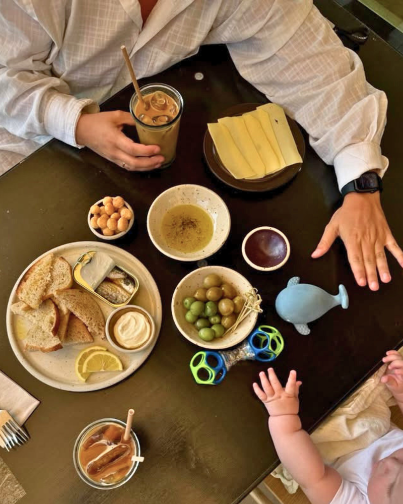 Et morgenbord med skiveskåret brød, oliven, kikærter, ost, dip, sardiner, citron, iskaffe, et blåhvalslegetøj og en babyhånd, der rækker op ved siden af en voksen med et ur på.