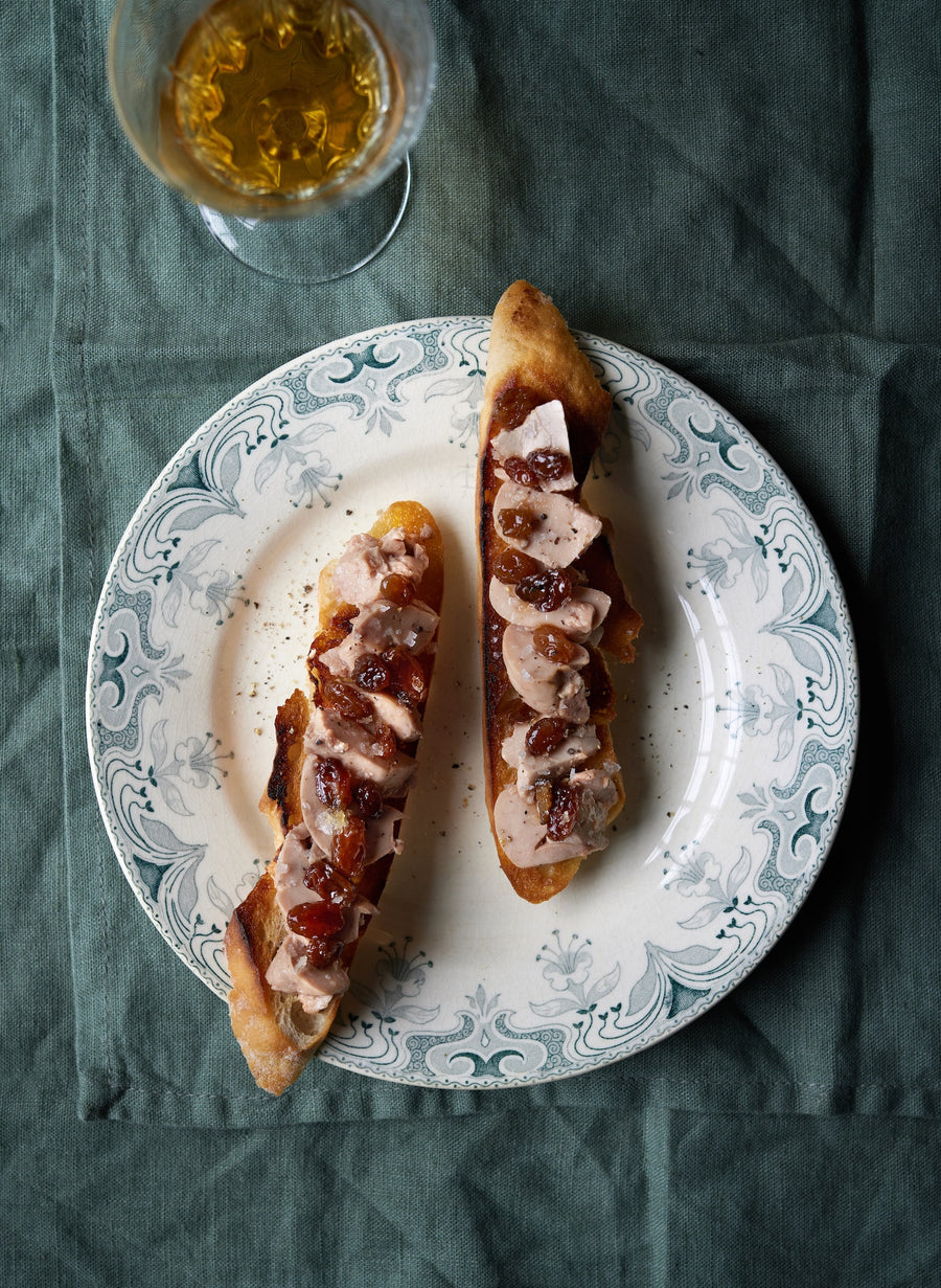 To skiver ristet baguette med kød og chutney er anrettet på en dekorativ tallerken med et glas ravfarvet drik over sig, alt sammen på en grøn dug.
