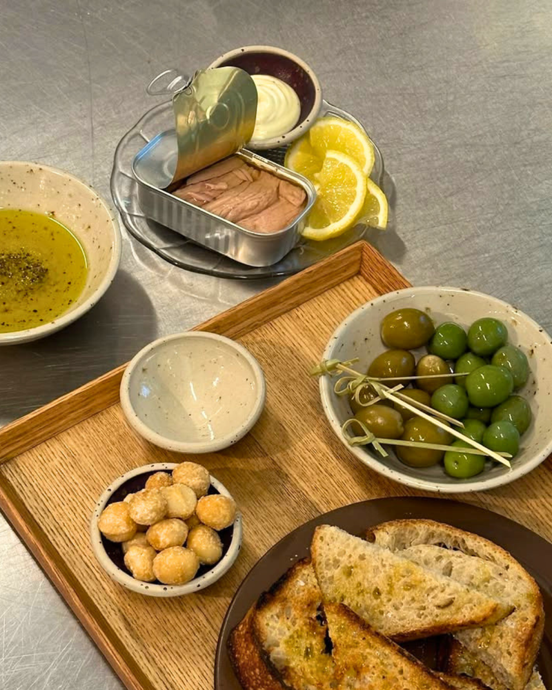 En træbakke med skåle med grønne oliven, macadamianødder, olivenolie og ristet brød. I nærheden står en åben dåse tun med citronbåde og saucer på en disk.