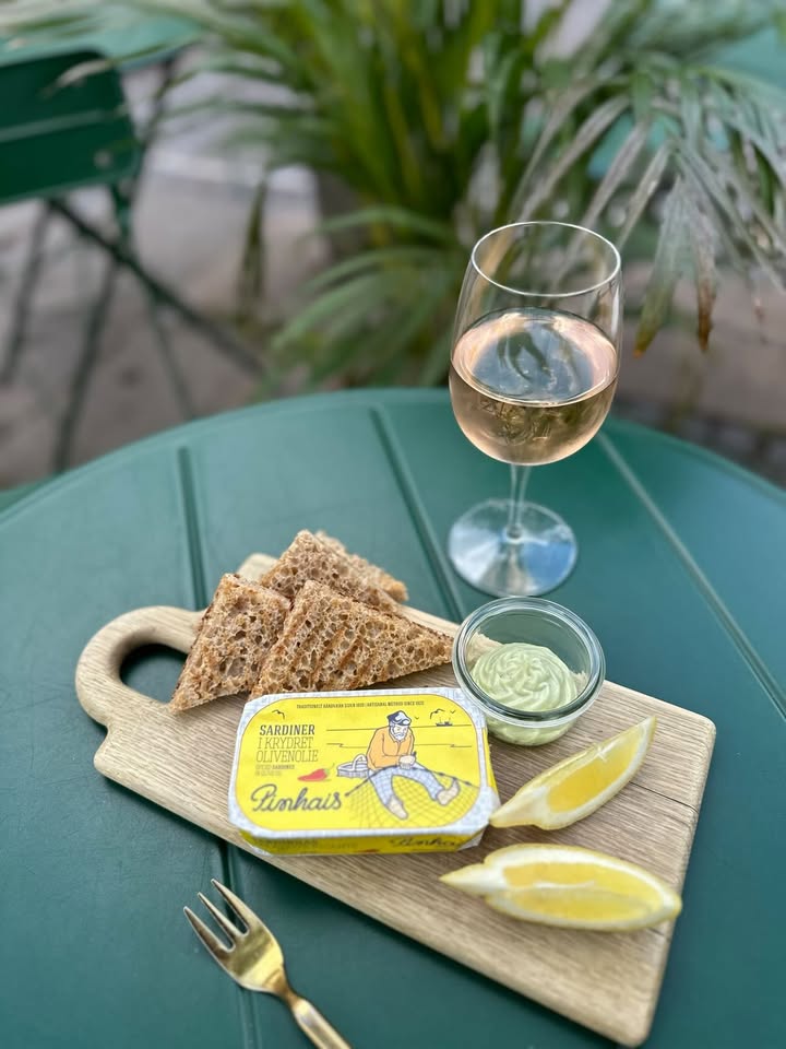 Et grønt bord med fuldkornsbrødskiver, citronbåde og en dåse sardiner på en træplade, ved siden af en lille skål med grønt pålæg og et glas rosévin. En potteplante står i baggrunden.