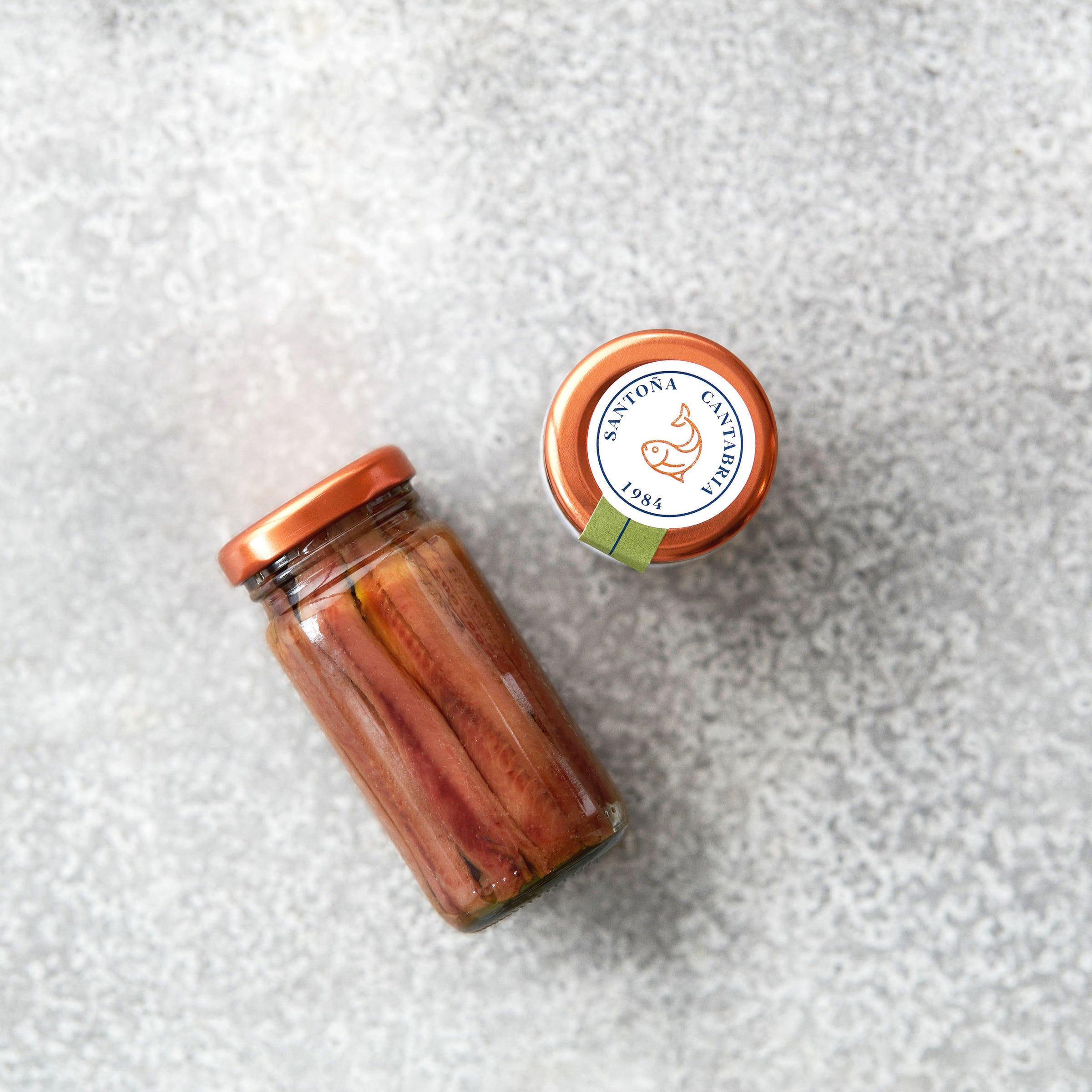 En glaskrukke med Pujado Solano OUTLET | 3 x Saltede ansjosfileter i solsikke med et kobberfarvet "SAVONA CENTOVENTI 1984"-låg, vist på en lysegrå struktureret overflade - rig på omega-3-fedtsyrer.