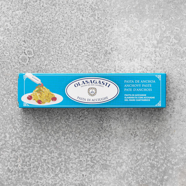 En blå rektangulær æske med Olasagasti Ansjospuré med olivenolie (60 g) ligger på en struktureret grå overflade og viser pasta med velsmagende ansjoscreme, tomater og flersproget information om bæredygtige ansjoser.