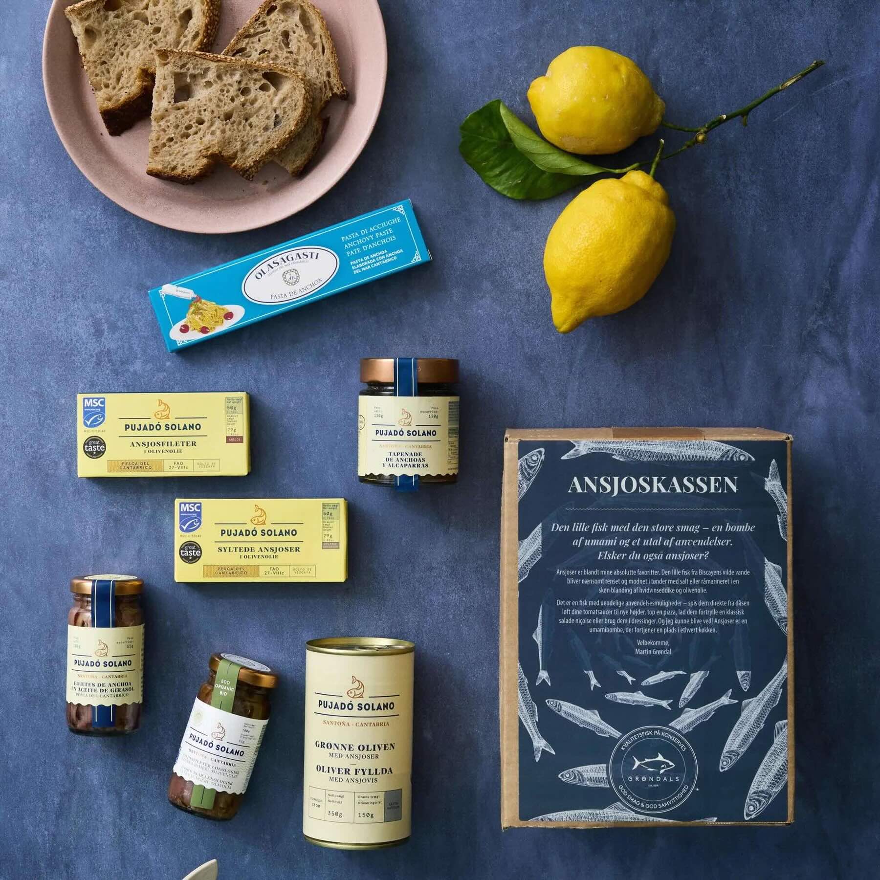 Et udvalg af middelhavsmad - herunder brød, citroner, glas med Grøndals konserverede fisk og skaldyr, en pakke ansjoser og Grøndals Ansjoskassen - er arrangeret på en blå flade.