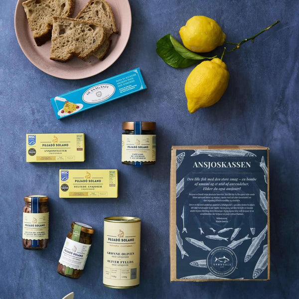 Et udvalg af middelhavsmad - herunder brød, citroner, glas med Grøndals konserverede fisk og skaldyr, en pakke ansjoser og Grøndals Ansjoskassen - er arrangeret på en blå flade.