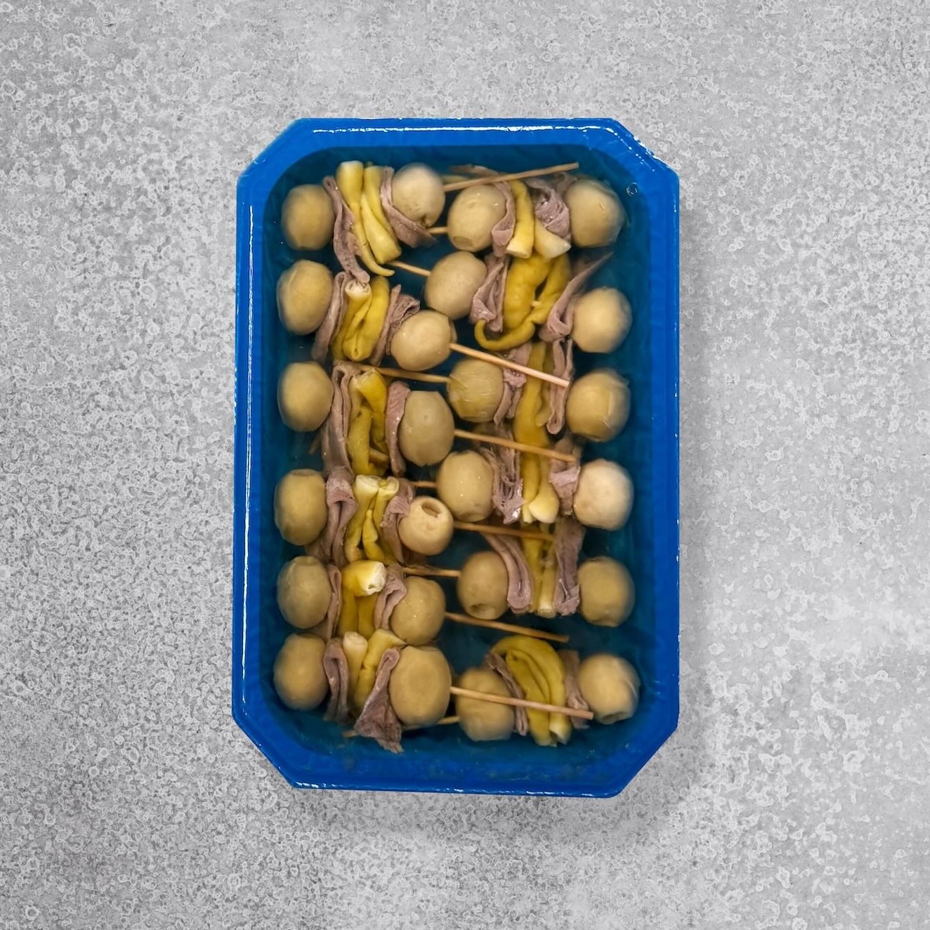 En blå bakke med Gildas spyd med saltede ansjoser fra Pujado Solano - spyd af manzanilla-oliven og gule syltede peberfrugter - smukt arrangeret på en grå overflade, fanger hjertet af klassiske spanske tapas og pintxos.