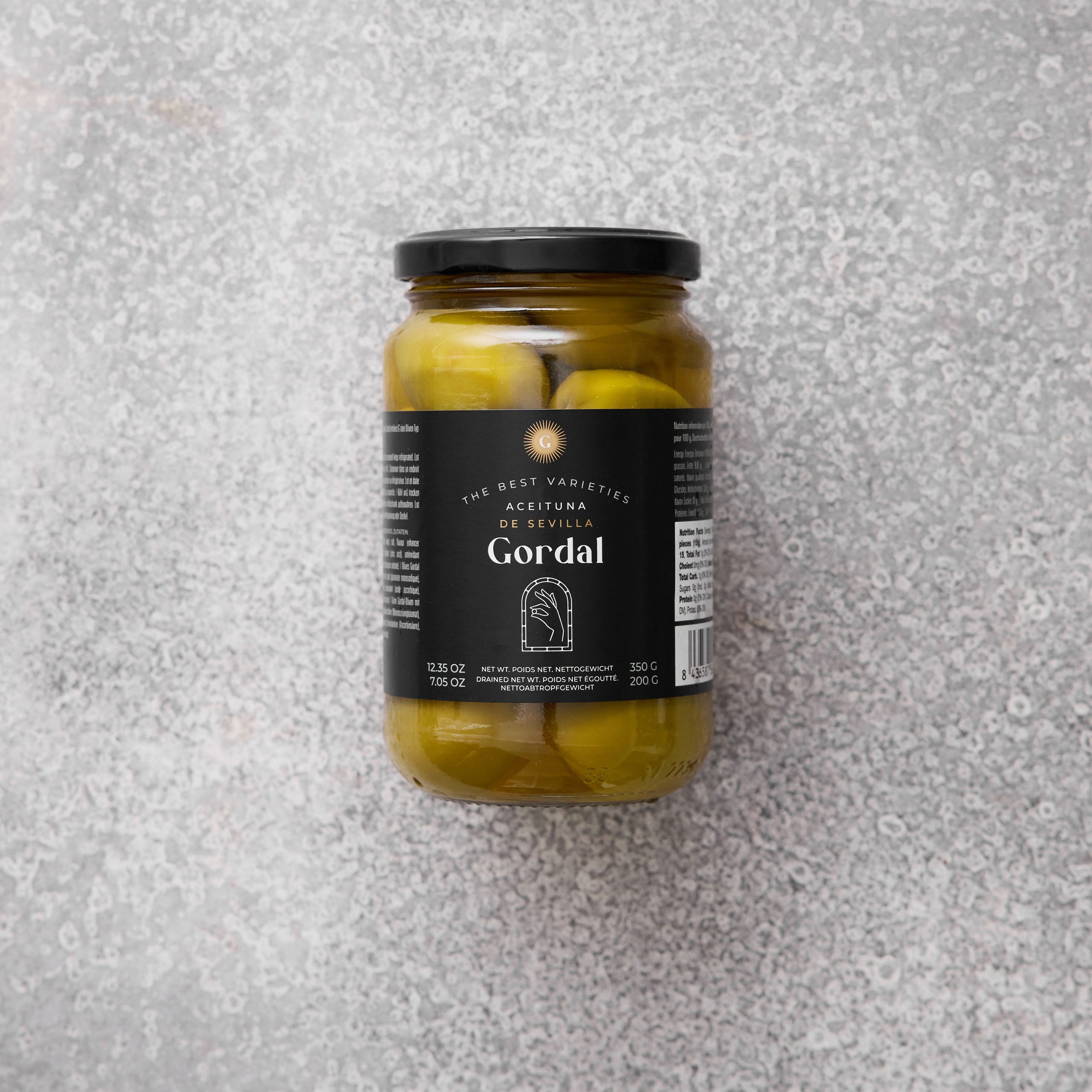 Glaskrukke med Gordal oliven fra Grøndals, med en sort etiket, set mod en grå tekstureret baggrund.