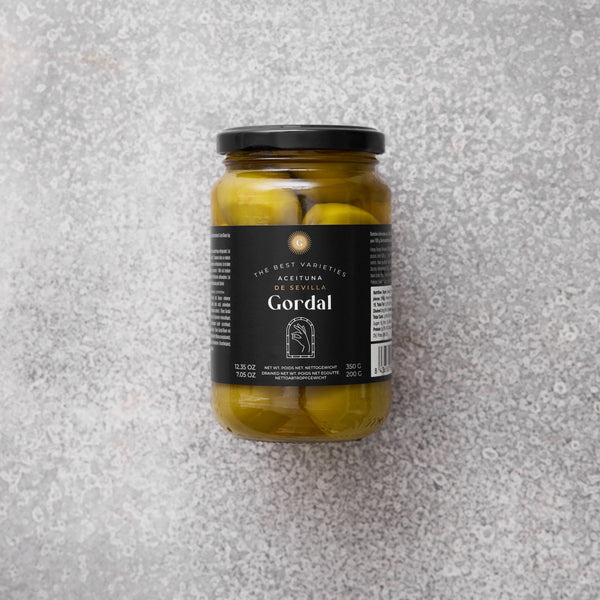 Glaskrukke med Gordal oliven fra Grøndals, med en sort etiket, set mod en grå tekstureret baggrund.