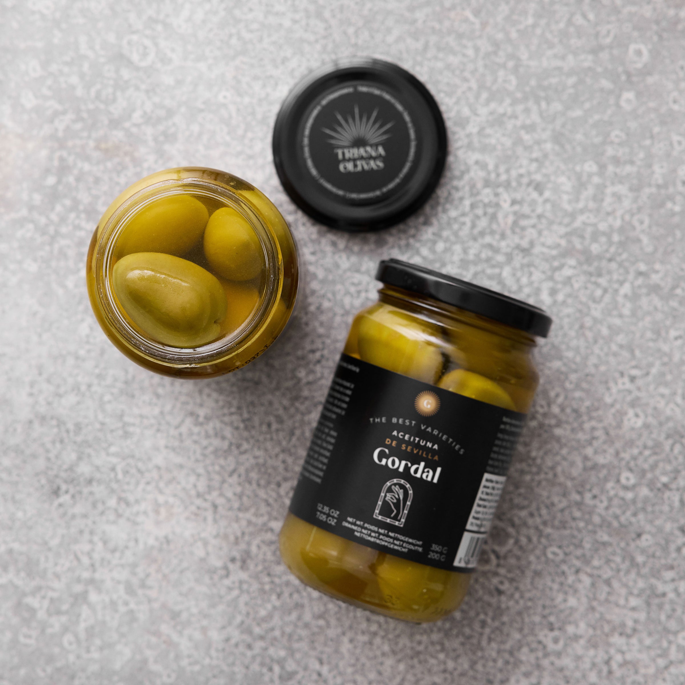 To glaskrukker med Gordal oliven fra Grøndals, hvor den ene er åben og set ovenfra, placeret på en grå tekstureret baggrund. Låget med mærket Triana Olivas er også synligt.