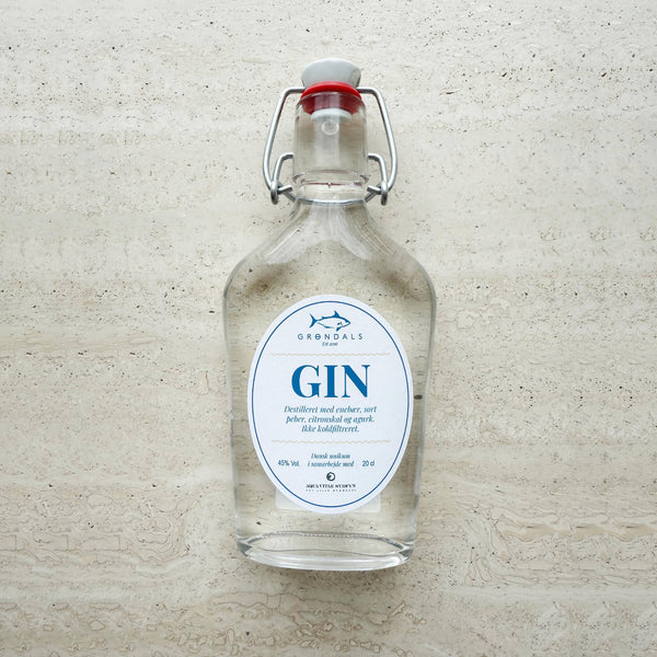 En klar Grøndals Gin - Frisk, krydret og skabt til fisk-flaske med svingprop og hvidblå etiket hviler på en tekstureret sten - en perfekt håndlavet gin til fisk fra Grøndals.