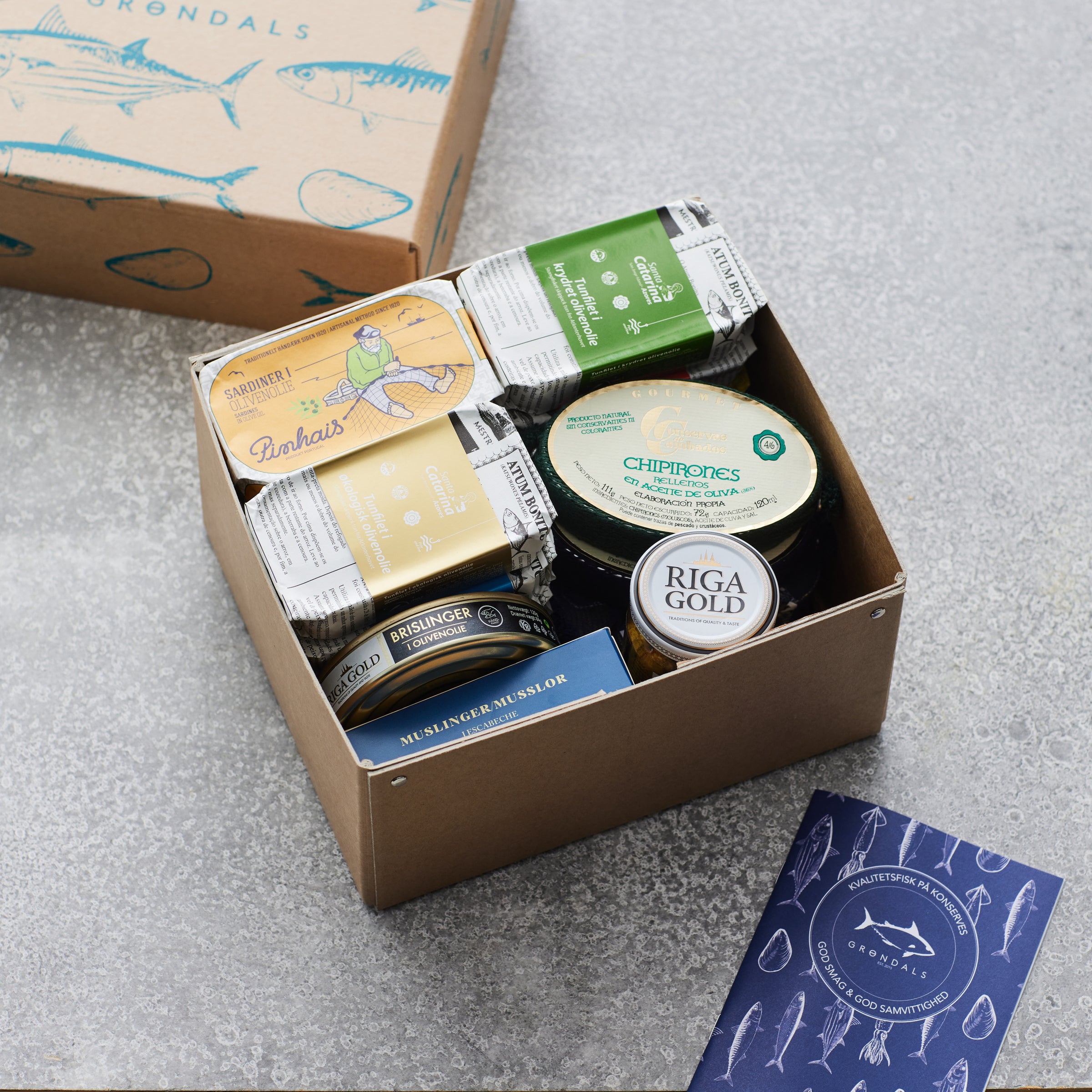 Grøndals Smagekasse fra Grøndals indeholder diverse fiskekonserves, herunder dåser med farverige etiketter og en Riga Gold-krukke. Et illustreret hæfte og en Grøndals-æske med et fiskemotiv er udstillet på en grå overflade.