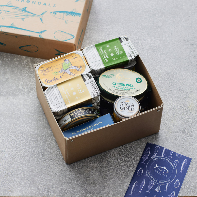 Grøndals Smagekasse fra Grøndals indeholder diverse fiskekonserves, herunder dåser med farverige etiketter og en Riga Gold-krukke. Et illustreret hæfte og en Grøndals-æske med et fiskemotiv er udstillet på en grå overflade.