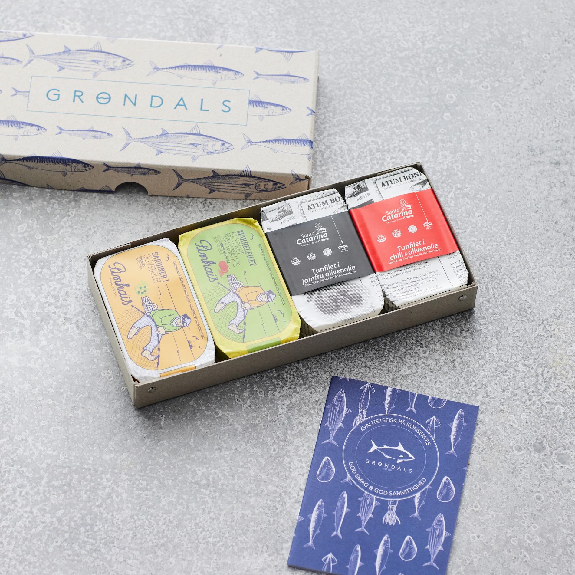 En åben Grøndals Værtsgave-æske viser fem farverige dåser med bæredygtighed-fokuseret konserves på en grå overflade ved siden af et blåt kort med fiskeillustrationer og Grøndals-logoet. Det ydre låg med fiskemønstre er placeret bagved.