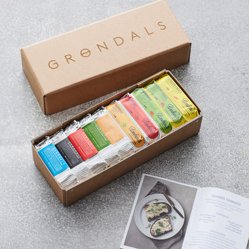 En brun Grøndals gavekasse fra Grøndals indeholder assorterede farverige dåser med dåsefisk, pænt arrangeret i den åbne æske. Et åbent opskriftshæfte med et foto af en fiskeret ligger i nærheden på en grå overflade.