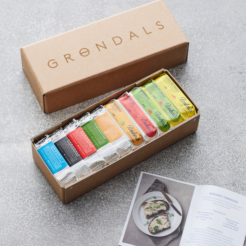 En brun Grøndals gavekasse fra Grøndals indeholder assorterede farverige dåser med dåsefisk, pænt arrangeret i den åbne æske. Et åbent opskriftshæfte med et foto af en fiskeret ligger i nærheden på en grå overflade.