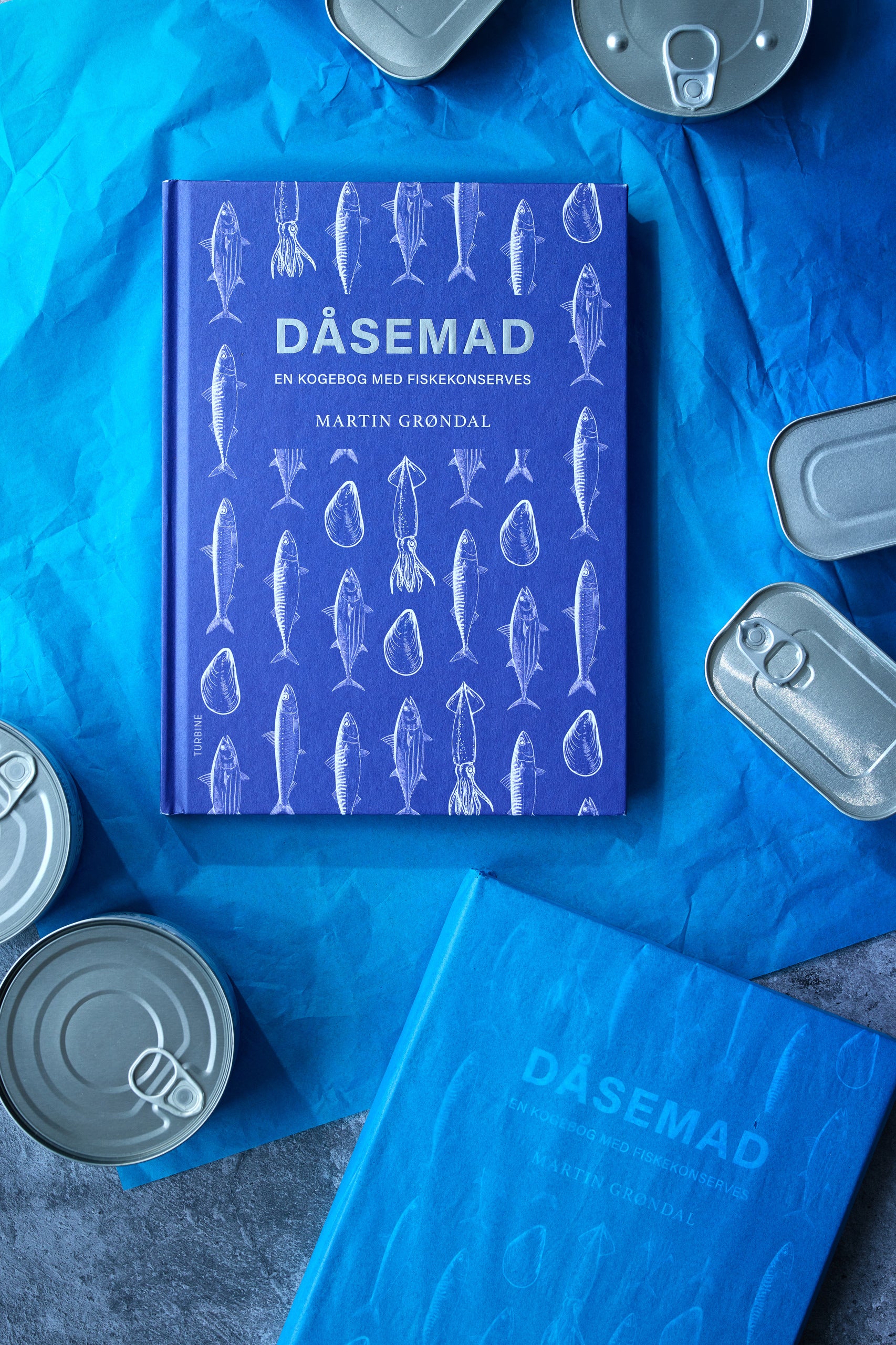Grøndals' Kogebogen DÅSEMAD, med et blåt omslag med fiskeillustrationer, hviler på blåt papir blandt uåbnede fiskekonserves. Et andet eksemplar af kogebogen er delvist synligt nederst.