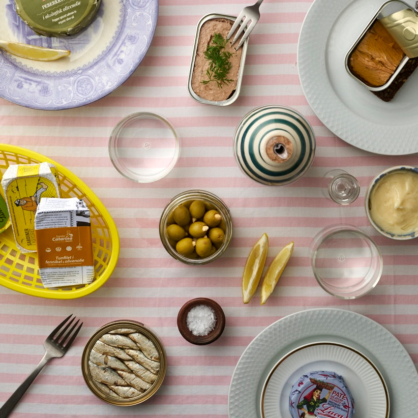 Et bord dækket på en lyserød og hvidstribet dug med tallerkener med fiskekonserves, grønne oliven, citronbåde, sennep, vandglas, salt og kiks i en gul kurv. En peberkværn står i midten.