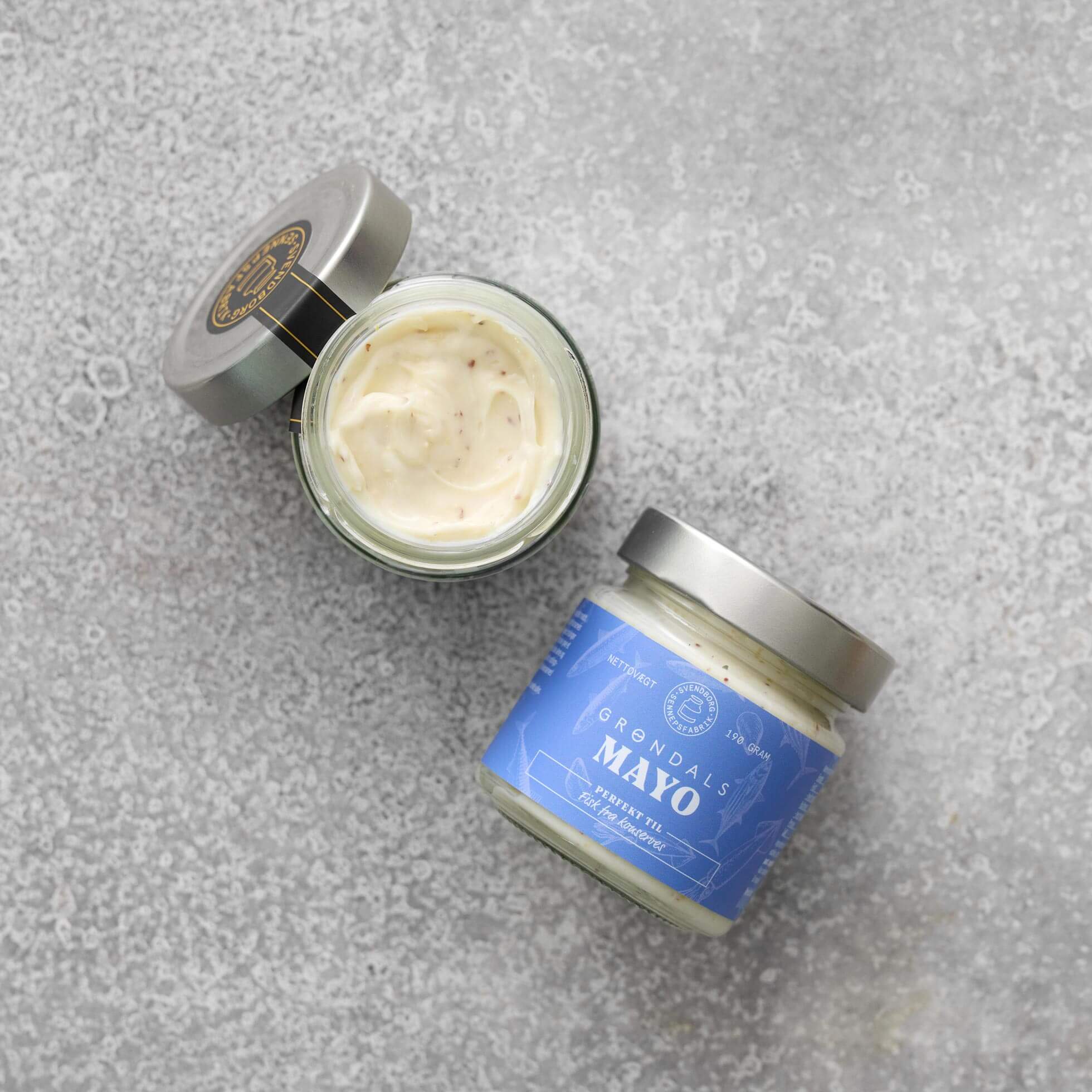 To glas Grøndals Mayonnaise – perfekt til fisk fra Grøndals, det ene åbent og afslører cremet mayonnaise med frilandsæg og det andet lukket med en blå etiket, står på en lysegrå tekstureret overflade – ideelt til servering med fiskekonserves.