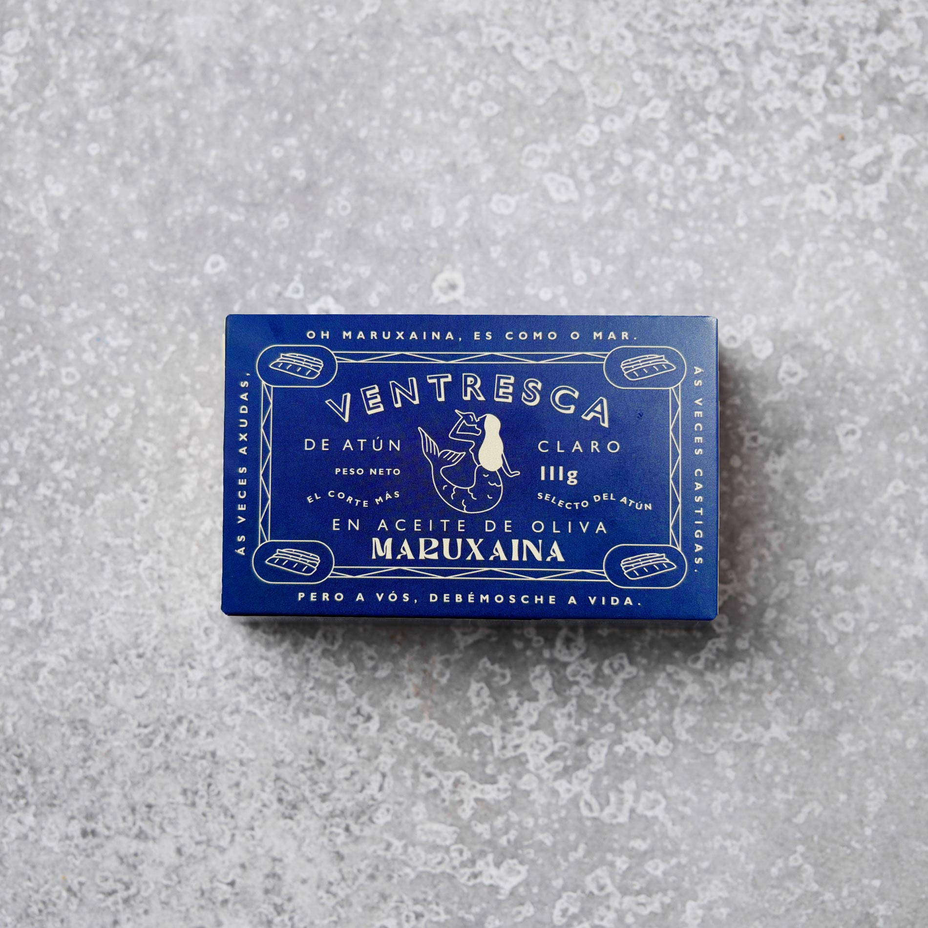 En blå rektangulær dåse med Don Gastronom Tun ventresca i olivenolie hviler på en spættet grå overflade med hvid tekst og en illustration af en kvinde med fisk på emballagen.