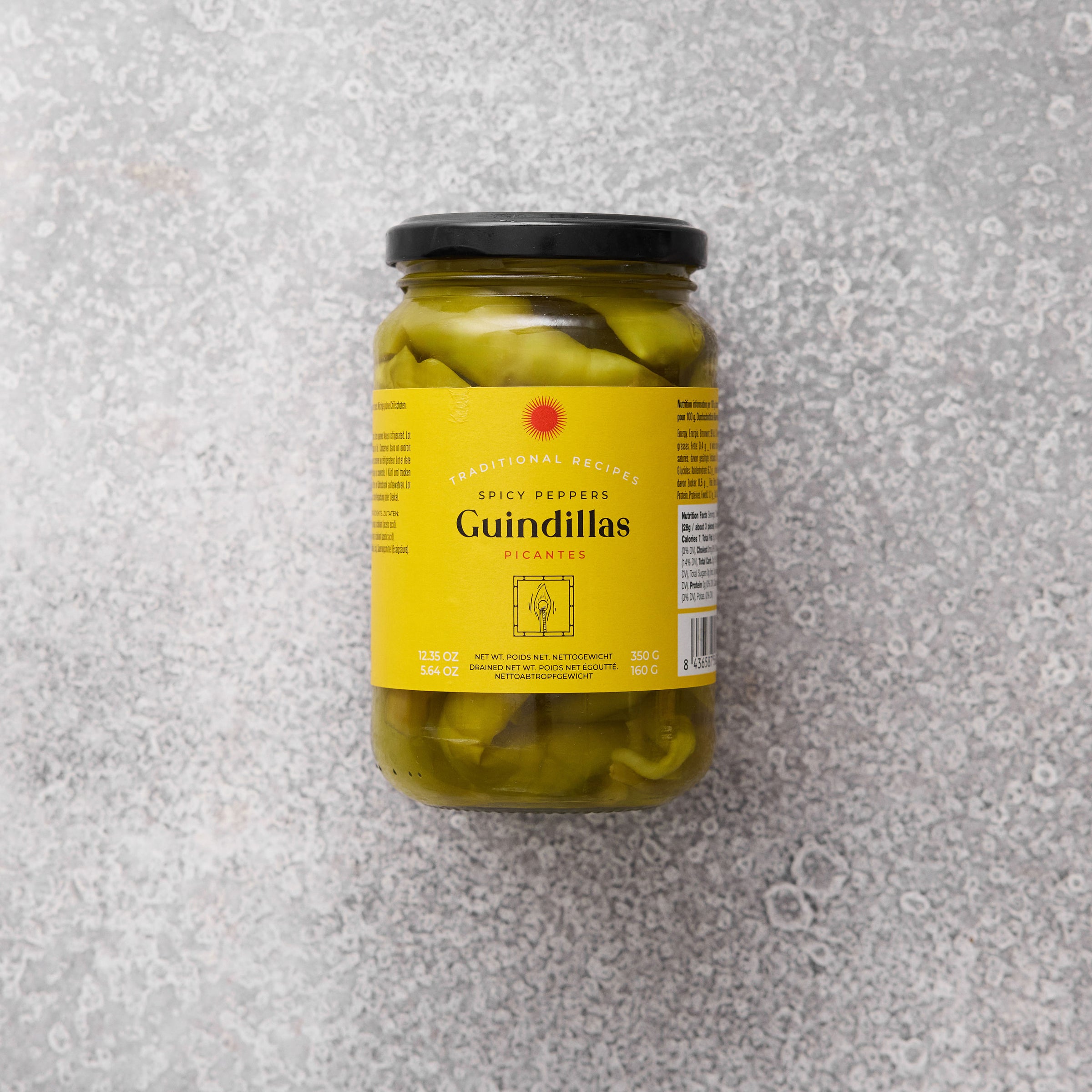 En glaskrukke med Grøndals Spicy guindillas peber med gul etiket og sort låg hviler på en struktureret grå overflade. Den tosprogede etiket antyder, at de er ideelle til tapas.