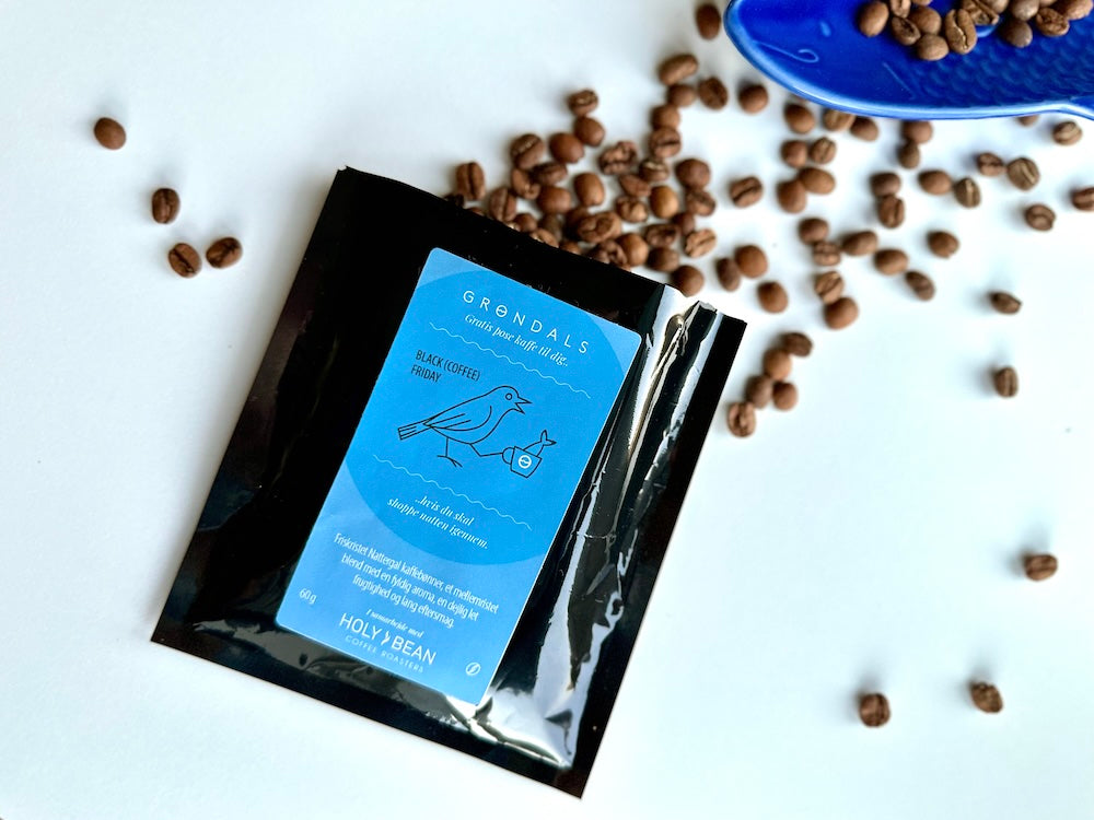 En sort kaffepakke med en blå etiket, der viser en fugl og et kaffekrus, ligger på en hvid flade. I baggrunden ses spredte kaffebønner og en del af et blåt fad med bønner.