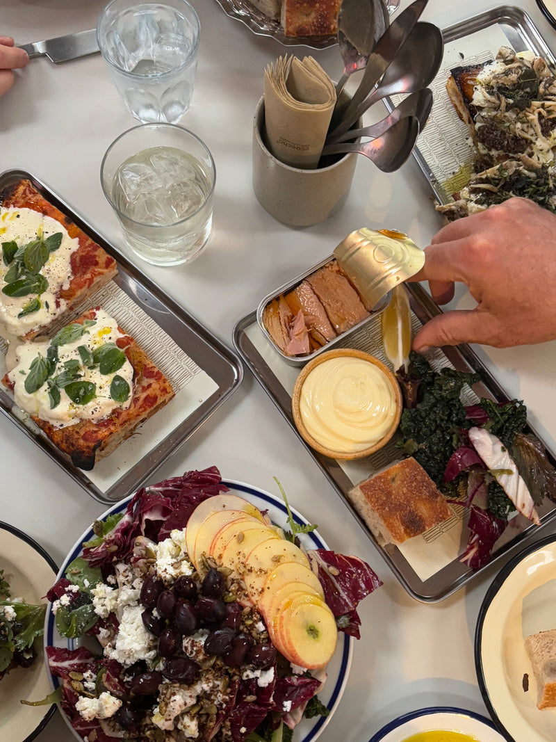 Et bord dækket med tallerkener med salat, æbleskiver, lasagne, brød, fisk på dåse med et låg, der er ved at blive åbnet, et glas vand og en beholder med servietter og skeer.