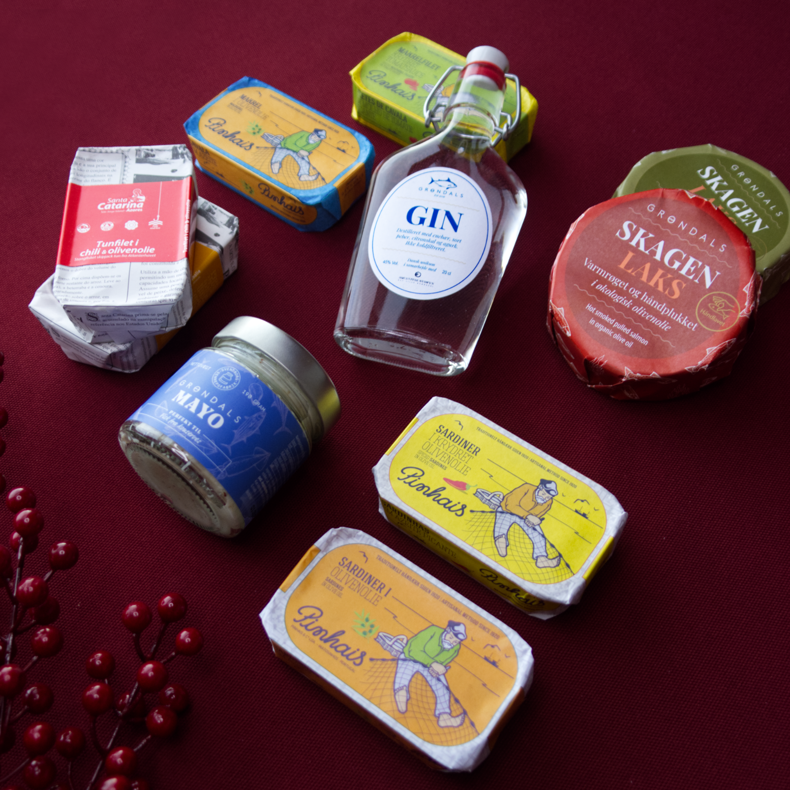 Grøndals Julekassen 2025 byder på fisk og skaldyr på dåse, varmrøget laks, mayo, gin, ost og festlige røde bær på en rød dug - en ideel julegave.