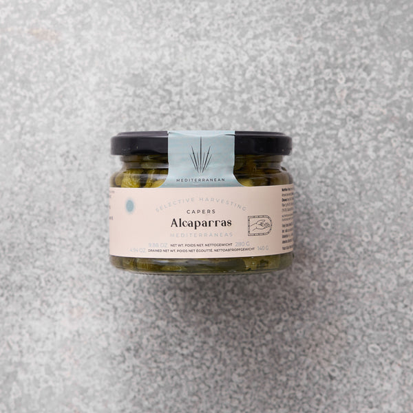 Glaskrukke med Alcaparras kapers fra Grøndals, med en lyseblå og beige etiket, set mod en grå tekstureret baggrund.