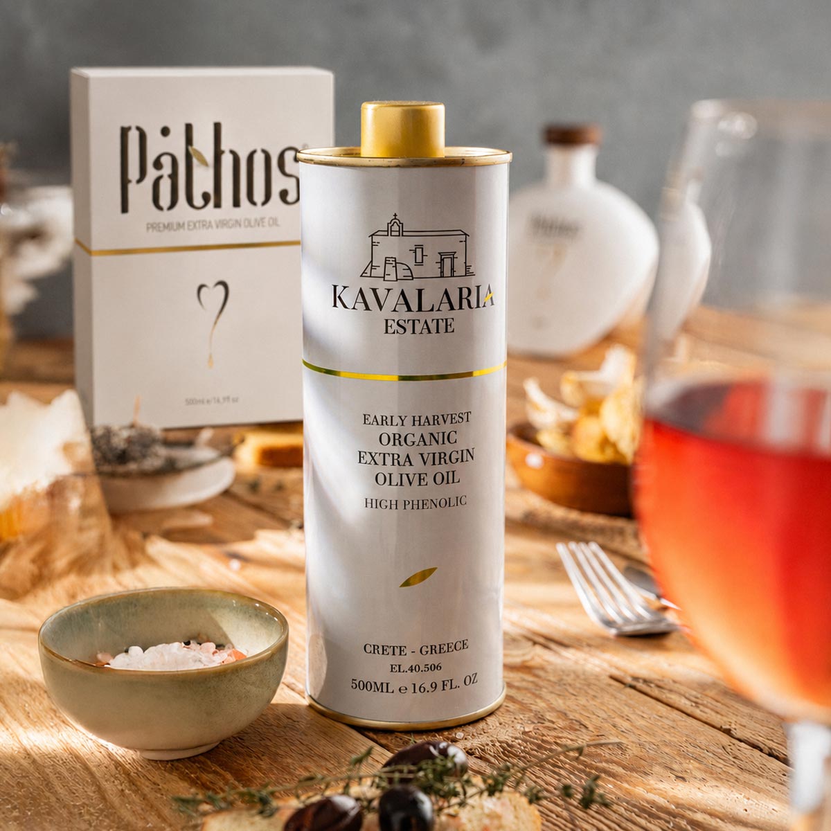 En dåse KAVALARIA ESTATE Økologisk Ekstra Jomfru Olivenolie står på et bord sammen med et glas rosé, snacks og en Pathos-olivenoliekasse i baggrunden.