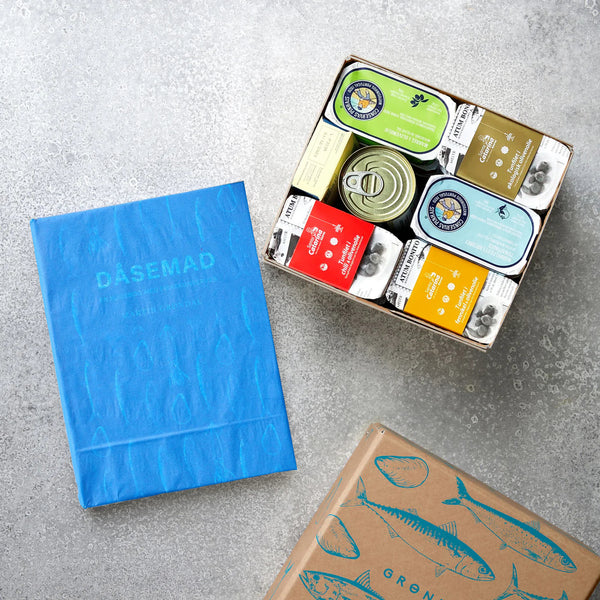 Grøndals' Smagekassen og DÅSEMAD - med en blå kogebog, en æske med forskellige fiskekonserves og en brun æske med blå fiskeillustrationer - vises på en grå flade.