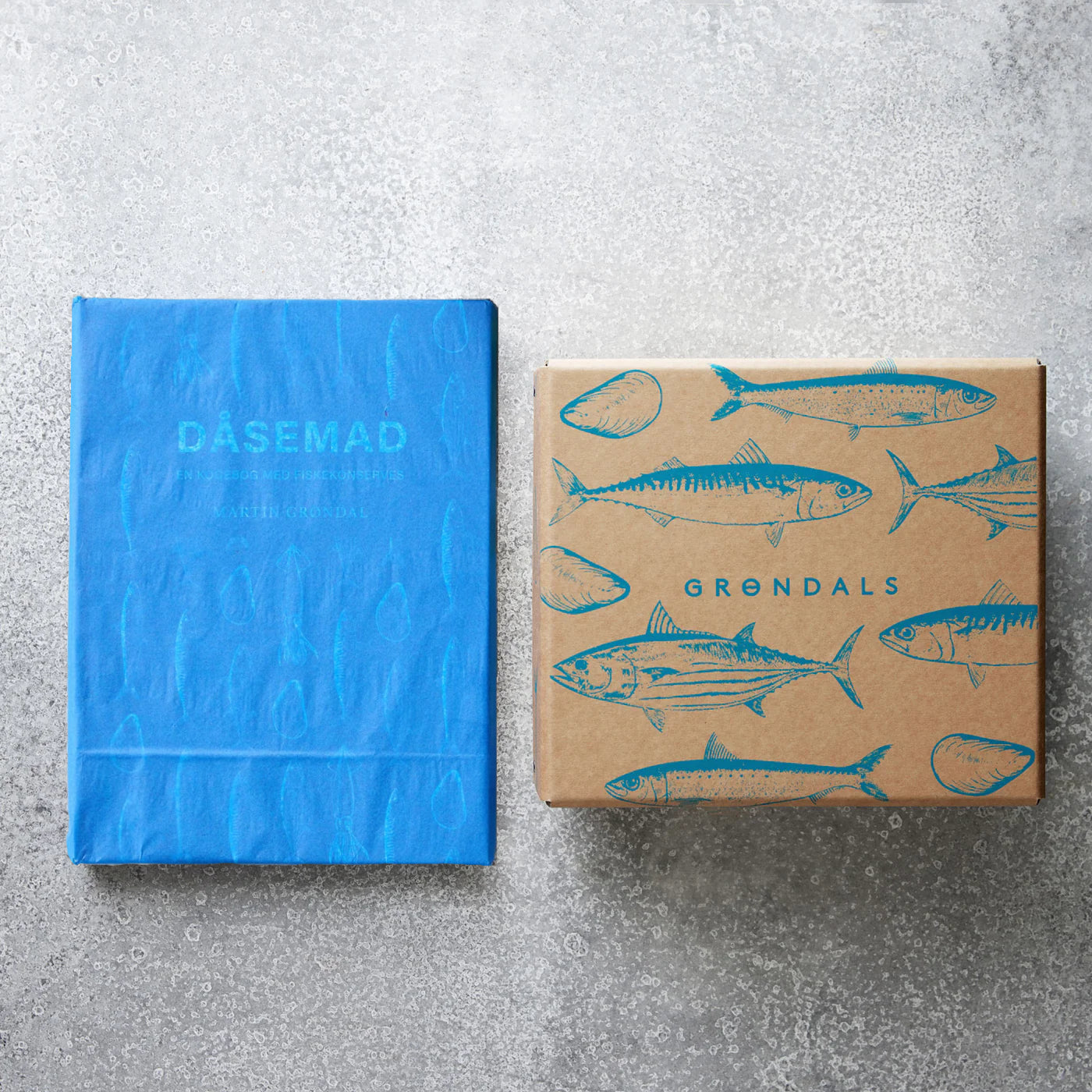 Grøndals Smagekassen og DÅSEMAD - en blå kogebog og en brun æske med blå fiskekonservesillustrationer - er udstillet sammen på en grå struktureret overflade.