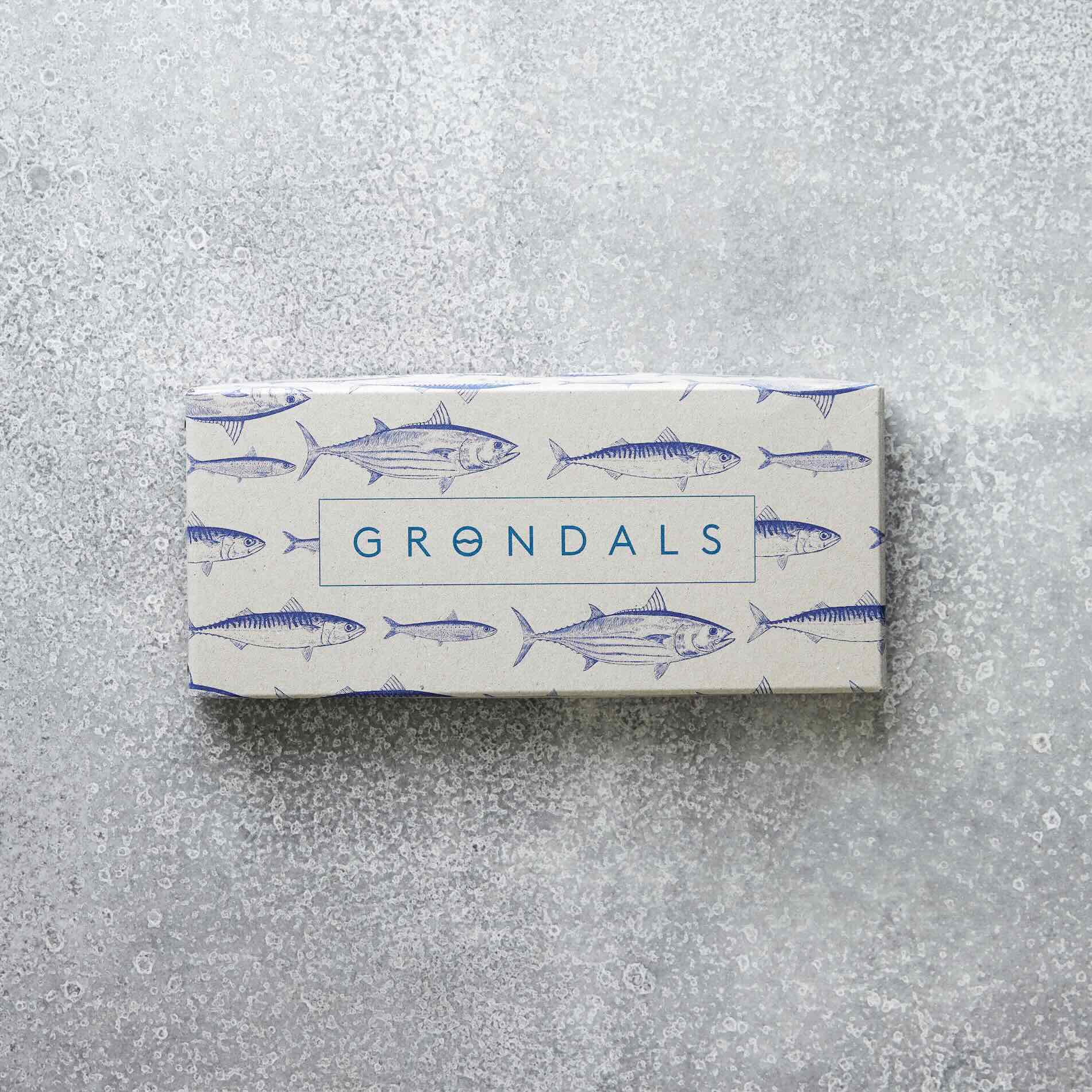 En rektangulær æske mærket "Æske til Værtsgave" fra Grøndals Praktik med et fiskemønster og "GRØNDALS" i blåt, placeret på en struktureret grå overflade.