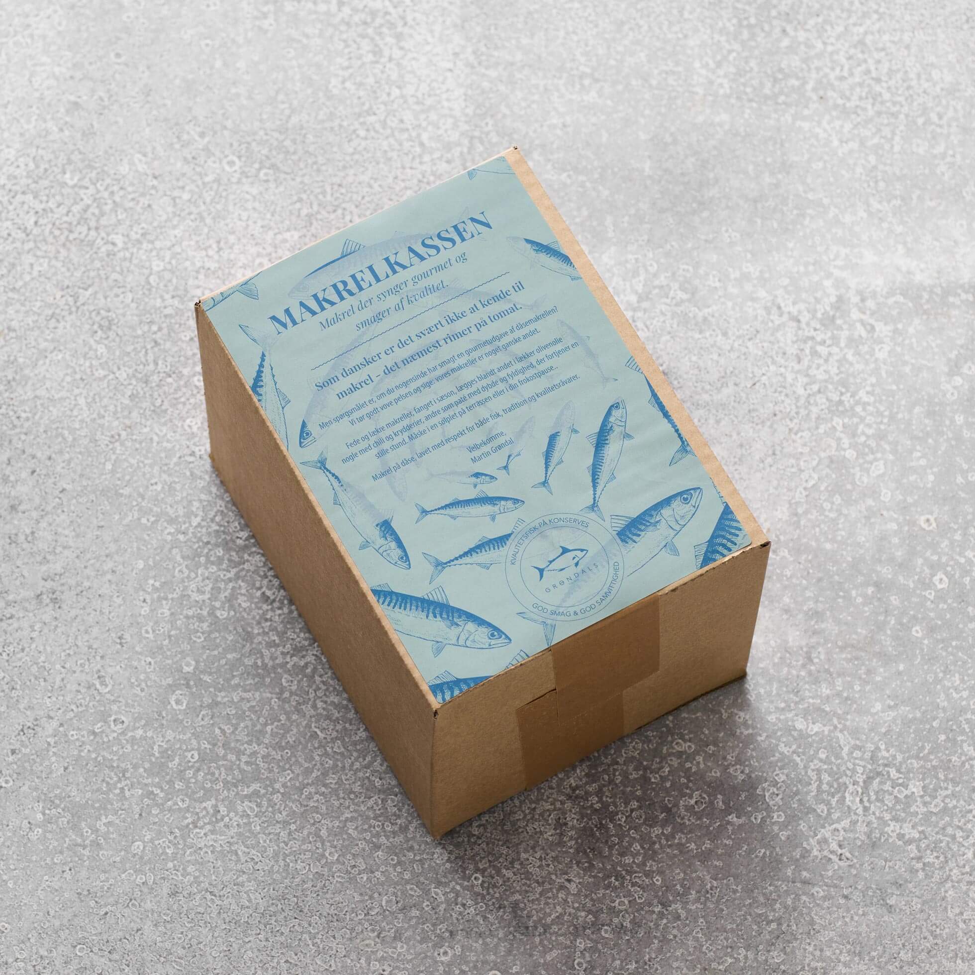 En Grøndals Makrelkassen papæske hviler på en grå tekstureret overflade, toppet med en blå etiket med illustrationer af makrol og tomat samt dansk tekst.