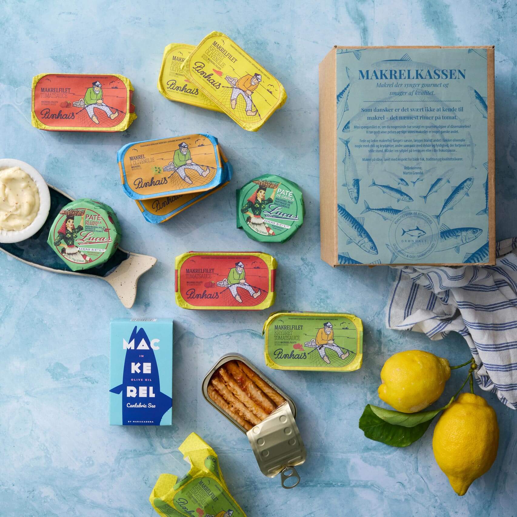 Et fladt lag farverig dåsefisk med Grøndals Makrelkassen, dåsemakrel i tomat, to citroner, et blåt makrelkort, sauce og en åben dåse fisk på en lyseblå overflade.