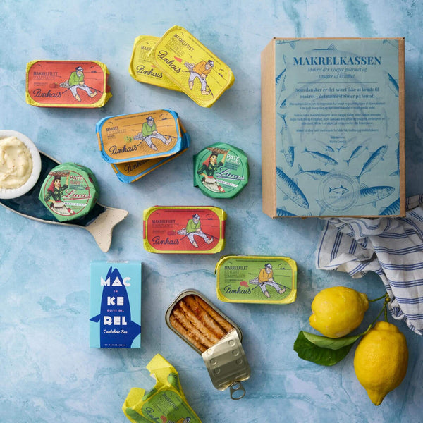 Et fladt lag farverig dåsefisk med Grøndals Makrelkassen, dåsemakrel i tomat, to citroner, et blåt makrelkort, sauce og en åben dåse fisk på en lyseblå overflade.